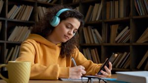 The photo depicts a young woman studying in a library. She's wearing headphones and has a phone in one hand and a pen in the other hand.